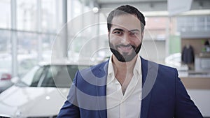 Portrait of successful Middle Eastern man posing in car dealership. Handsome young businessman looking back at new white