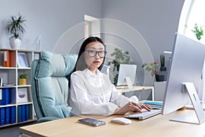 Portrait of successful office worker Asian woman, looking at camera smiling, business woman in glasses working and