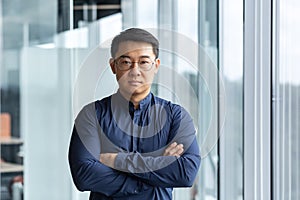 Portrait of successful and serious asian boss in shirt, man looking at camera with crossed arms inside modern office