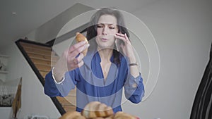 Portrait of successful serious caucasian woman choosing croissant and talking on the phone at home. Beautiful confident