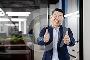 Portrait of a successful smiling Asian male businessman standing in a suit in an office center and showing the super