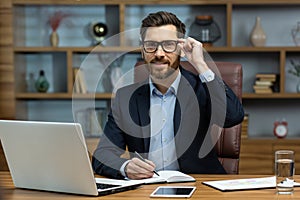 Portrait of successful smiling man in office, mature businessman looking at camera cheerfully, senior boss in glasses