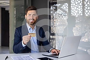 Portrait of successful and smiling young banker, office worker sitting at desk in office holding and showing credit card