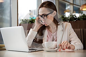 portrait of a successful young business woman with dark hair talking on the phone behind a laptop at a table in