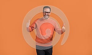 Portrait of successful young man in eyeglasses showing thumbs up signs on isolated orange background