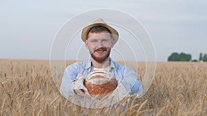 Portrait of sucessful agronomist, young man gives you freshly baked bread and smiles at camera in wheat crop field