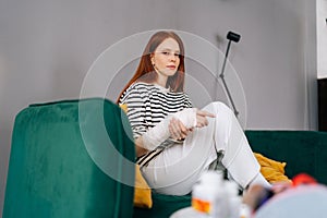 Portrait of suffering from pain unhappy young woman with broken right hand wrapped in white gypsum bandage sitting on