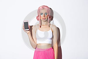 Portrait of sulking girl complaining and showing something disappointing on mobile phone screen, standing in pink hair