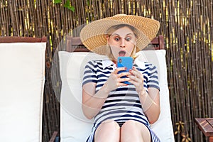 Portrait of suprised young adult blogger woman in hat and dress is sitting on sunbed and holding phone, looking at screen reading