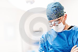 Portrait of surgeon, cosmetic plastician looking down with surgical lamps in background. Doctor with loupes