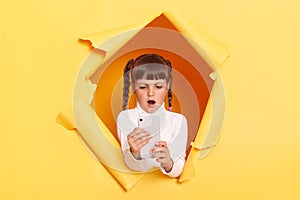 Portrait of surprised amazed shocked little girl with braids wearing white turtleneck, holding smart phone, looking at display,