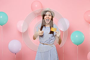 Portrait of surprised amazed woman in straw summer hat blue dress holding credit card showing thumb up on pink