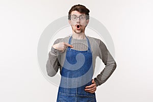 Portrait of surprised and amazed young caucasian guy in apron pointing at himself and looking disbelief, gasping drop