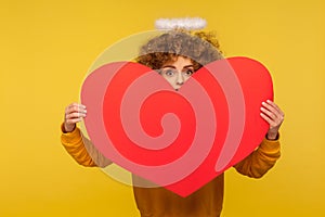 Portrait of surprised angelic curly-haired woman with saint nimbus peeking out of big red heart, yellow background