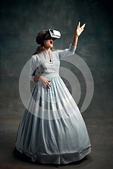 Portrait of surprised aristocratic woman wearing blue historical dress and VR glasses waving hand in air. Gadgets. Queen