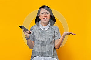 Portrait of surprised asian woman holding mobile phone with confused and unhappy expression on yellow background