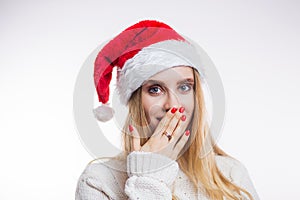 Portrait of surprised attractive young woman in red Santa hat and beige sweater on white background. A girl with a bright manicure