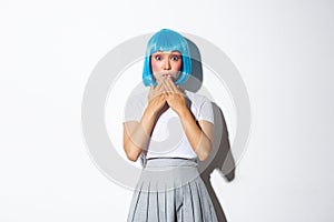 Portrait of surprised beautiful asian girl in glamour blue wig, looking amazed, holding hands near lips thrilled