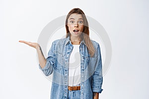 Portrait of surprised beautiful girl holding in hand, spread hand sideways and showing object in palm and smiling amazed