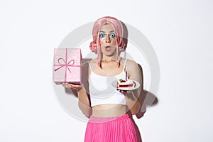 Portrait of surprised beautiful girl in pink wig, receive birthday gift, holding b-day cake and smiling happy, standing