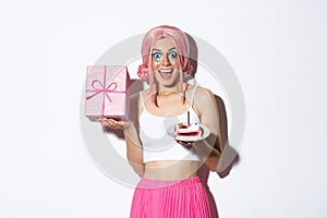 Portrait of surprised beautiful girl in pink wig, receive birthday gift, holding b-day cake and smiling happy, standing