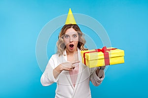 Portrait of surprised beautiful woman with birthday party cone on head pointing at present box and looking with amazement. blue