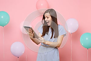 Portrait of surprised beautiful woman wearing blue dress holding using tablet pc computer on pastel pink background with