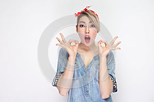 Portrait of surprised beautiful young woman in casual blue denim shirt with makeup and red headband standing with ok sign and