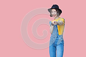 Portrait of surprised beautiful young woman in yellow t-shirt, blue denim overalls with makeup and black hat standing, amazed,