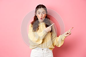 Portrait of surprised brunette girl gasping impressed, pointing fingers right at copy space aside, showing way to logo