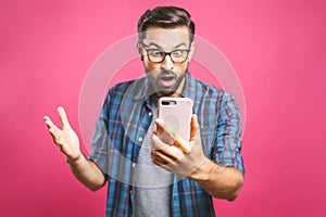 Portrait of a surprised casual man looking at mobile phone isolated over pink background