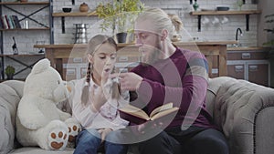 Portrait of surprised Caucasian girl listening to father reading her book. Positive man touching daughter`s nose with