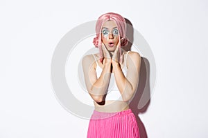 Portrait of surprised caucasian girl in pink wig and bright makeup, gasping amazed and stare at camera in awe, standing