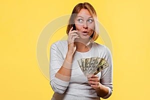 Portrait of a surprised caucasian woman holds a fan of dollars and talks on the phone. Yellow background. The concept of discounts