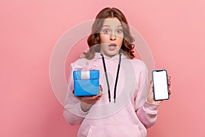 Portrait of surprised curly-haired teenage girl in hoodie holding smartphone and wrapped gift box, smiling to camera