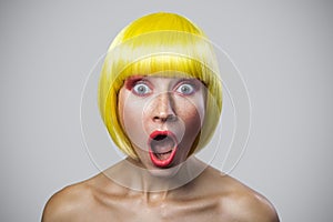 Portrait of surprised cute young woman with freckles, red makeup and yellow wig, looking at camera with amazed wonder face