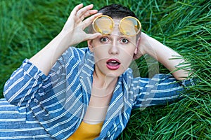 Portrait of surprised cute young woman with short hair in casual blue striped suit lying down on green grass, hoding yellow photo