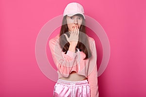 Portrait of surprised European brunette teenager covers her opened mouth with hand, dressed in stylish rose sweatpants, hoodie and