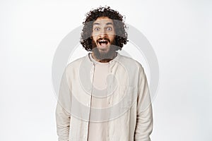 Portrait of surprised and excited middle eastern man, drop jaw and gasping in awe, looking at something amazing, white