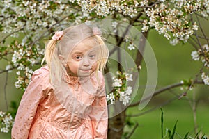 Portrait of surprised or frustrated little girl outside near flowering tree with funny grimace on her face. Spring day