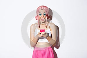 Portrait of surprised girl looking at happy news on mobile phone, wearing pink wig for party, halloween celebration