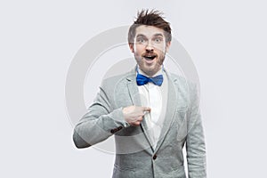 Portrait of surprised handsome bearded man in casual grey suit and blue bow tie standing, pointing himself and looking at camera