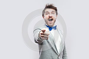 Portrait of surprised handsome bearded man in casual grey suit and blue bow tie standing pointing and looking at camera with