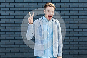 Portrait of surprised handsome young blonde man in casual style standing with victory or peace gesture and looking at camera with