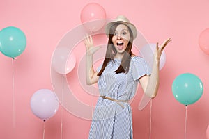 Portrait of surprised happy young woman wearing straw summer hat and blue dress spreading hands on pastel pink