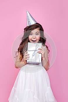 Portrait of surprised kid girl n birthday party princess hat holding gift box on a pink studio background