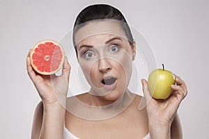 Portrait of surprised middle aged woman with perfect face skin holding fresh slice grapefruit and apple in her hands and open