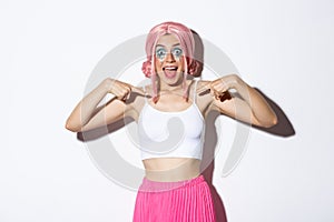 Portrait of surprised party girl with pink wig and bright makeup, pointing at herself and smiling excited, showing her