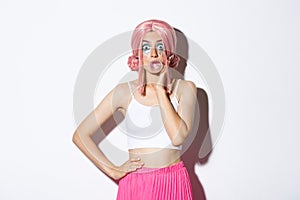 Portrait of surprised pretty girl in pink wig, blowing bubble gum and looking amazed at camera, standing over white