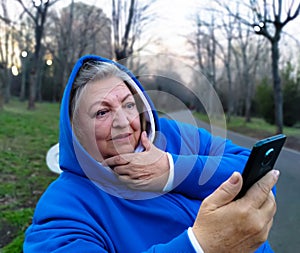 Portrait of surprised senior woman looking at mobile, sitting on bench in park at sunset. Close up